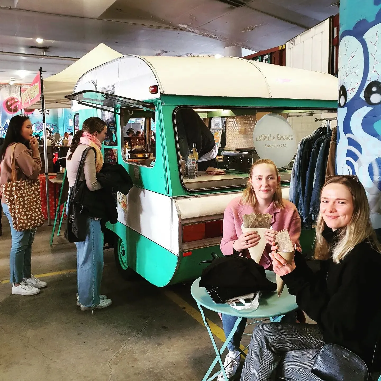 Two smiling customers holding crepes at a table beside the La Belle Epoque caravan in a market hall